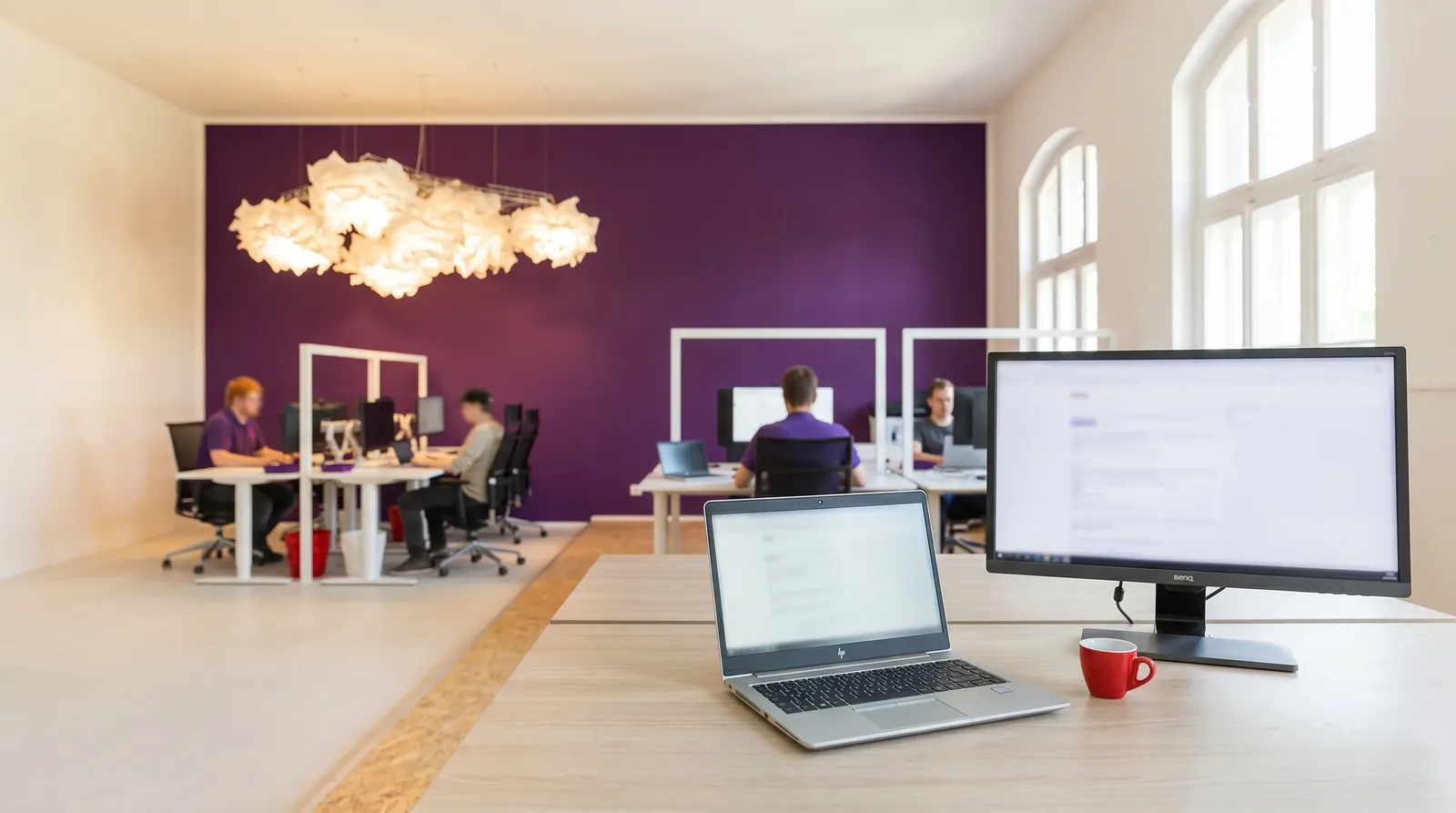 Heller moderner Arbeitsplatz mit HP-Notebook und BenQ-Monitor, Team im Hintergrund vor lila Akzentwand